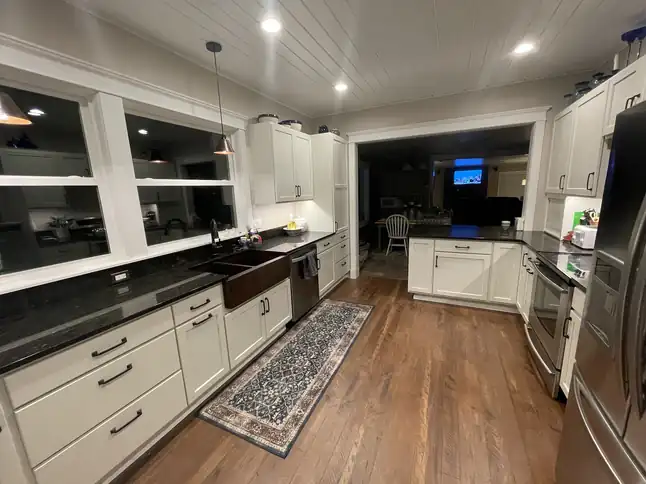 Wide view of a remodeled kitchen with white shaker cabinets, a dark farmhouse sink, hardwood floor and a patterned runner rug