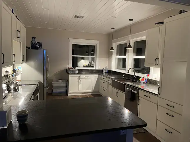 Remodeled kitchen at night with white cabinets, dark granite counters, a farmhouse sink under the window and two pendant lights over a peninsula