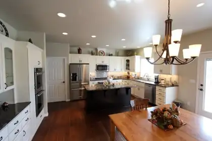 Large kitchen with white cabinets, a dark center island with granite top, double wall ovens, stainless appliances, dark wood floor and a bronze chandelier