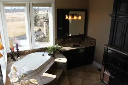 Master bath with a corner soaking tub under a large window looking out over open farmland, dark vanity with granite top and mosaic backsplash