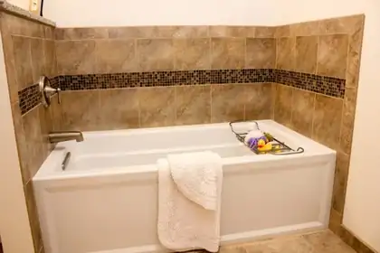 Built-in white soaking tub with a tan tile surround and a band of mosaic glass tile, folded white towel on the tub edge