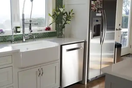 Farmhouse sink in a white kitchen with a green tile backsplash and a stainless refrigerator