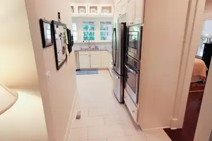 Long hallway leading to a white kitchen with a stainless refrigerator and wall ovens