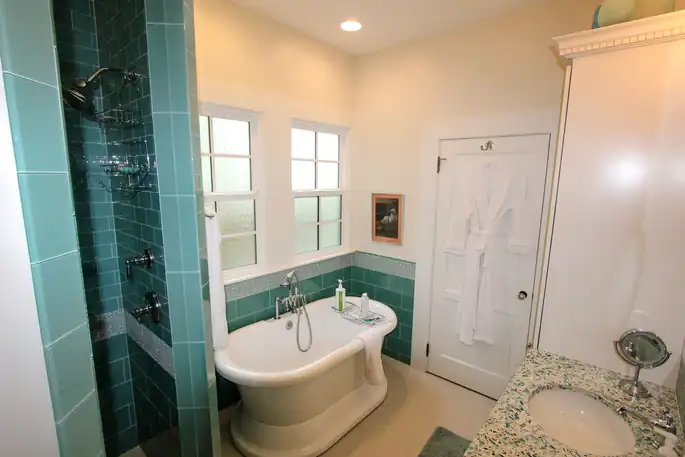 Remodeled bathroom with teal tile walk-in shower, a freestanding white tub with a floor-mounted faucet, a bathrobe on the door and a mosaic glass counter